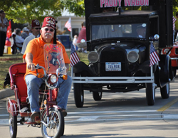 Man with fez in orange shirt on tricycle in front of a black car
